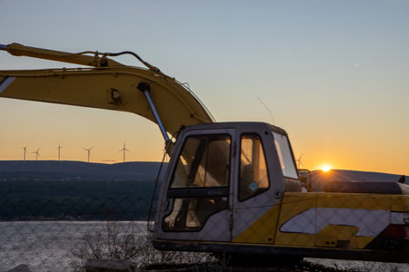 An excavator rests in the foreground at dusks light, with wind turbines silhouetted in the background, representing industrial progress and the drive towards renewable energy.の写真素材