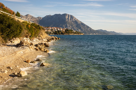 A stunning coastal scene showcases clear turquoise water gently lapping against rugged stones, framed by green foliage and majestic mountains in the background.の写真素材