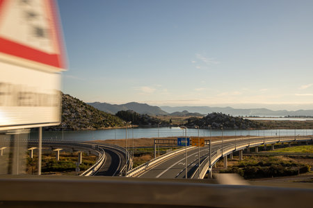 This image captures a winding road alongside tranquil water, framed by distant mountains and natural beauty, showcasing a perfect blend of nature and infrastructure.の写真素材