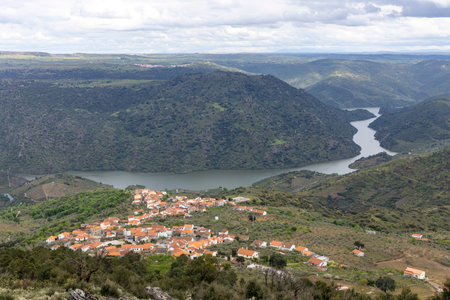 A picturesque village with red roofs nestled beside a serene river in a lush green landscape, under a cloudy sky, offering stunning views.の写真素材