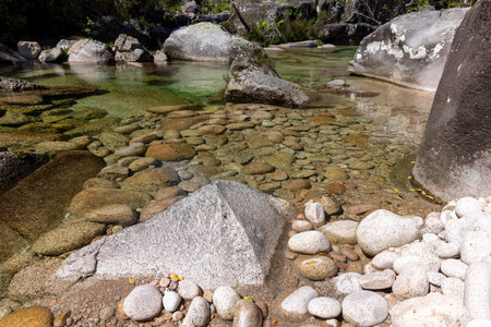 The image presents a high-angle, close-up view of a shallow stream bed.  The stream bed is predominantly composed of various sizes of rocks and pebbles, ranging from small, smooth stones to larger, angular rocks.  The sediment appears light beige/brownish, mixed with patches of darker material possibly indicating algae or organic matter.  The water is relatively clear, revealing the texture and composition of the stream bed.  The depth of the water is shallow, allowing for a clear view of the rocks beneath the surface.  The overall color palette is muted, with earth tones predominating.  A section of what appears to be a concrete or stone structure is partially visible in the upper right corner.  The lighting suggests natural daylight conditions. The focus is sharp on the rocks and sediment in the foreground, with a slight softening in focus as the image recedes to the background.の写真素材