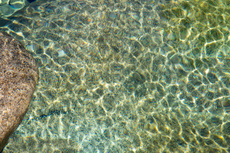 High-angle, close-up shot depicting the surface of shallow, clear water overlying a bed of smooth,...