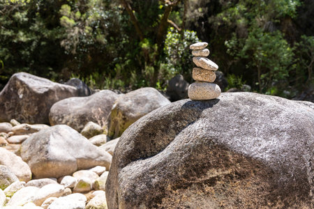 The image showcases a digitally captured photograph of a carefully balanced stack of smaller stones atop a larger, dark gray boulder. The boulder rests amidst a collection of other rocks and pebbles of varying sizes, suggesting a natural, possibly riverbed or forest floor setting. The background features blurred green foliage, indicating the presence of trees and suggesting a wooded area. The lighting suggests an outdoor scene bathed in bright, natural daylight, creating defined shadows and highlighting the textures of the rocks and stones. The overall color palette is composed of earth tones, primarily grays, browns, and greens.  The focus is sharp on the stacked stones and the foreground boulder, with depth of field subtly blurring the background elements.の写真素材