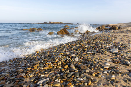 The image shows a section of a rocky seashore.  The foreground is dominated by a large expanse of dark-colored pebbles and small rocks covering the beach.  Small waves are breaking on the shoreの写真素材