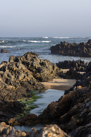 The image shows a close-up view of dark, rough-textured rocks on a coastline.  The rocks are irregularly shaped and appear weathered, showing signs of erosion.  Ocean waves are visible.の写真素材