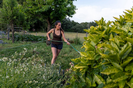 The image shows a woman, from the knees up, actively spraying plants in a garden setting.  She is wearing a dark-colored sleeveless top and dark shorts.の写真素材