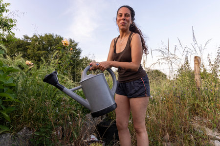 A low-angle, medium outdoor shot depicts a young woman with long dark hair smiling as she waters plants with a grey watering can. The woman wears a brown tank top and denim shorts.の写真素材