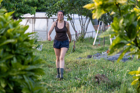 Eye-level medium shot of a young woman with brown hair walking through a garden. She is wearing a dark-colored tank top, dark-colored shorts, and dark-colored boots.の写真素材