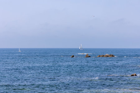 A sailboat is sailing in the ocean near some rocks. The sky is clear and the water is calm