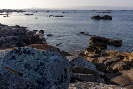 A rocky shoreline with a large rock in the foreground. The water is calm and the sky is a light blueの写真素材
