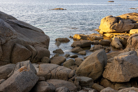 A rocky shoreline with a large rock in the foreground. The water is calm and the sky is a light blueの写真素材