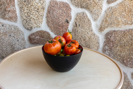 A bowl of tomatoes sits on a table. The bowl is black and the tomatoes are red. The bowl is placed on a wooden tableの写真素材