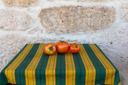 Three tomatoes are sitting on a table with a striped cloth underneath. The table is located in front of a stone wallの写真素材