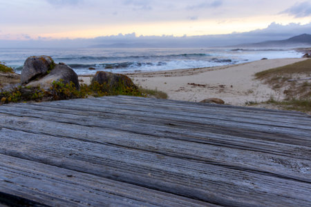 A wooden table sits on a beach near the ocean. The table is empty and the beach is rocky. The sky is...