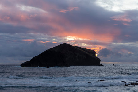 A rocky island is visible in the distance, with the sun setting behind it. The sky is filled with clouds, creating a moody atmosphereの写真素材