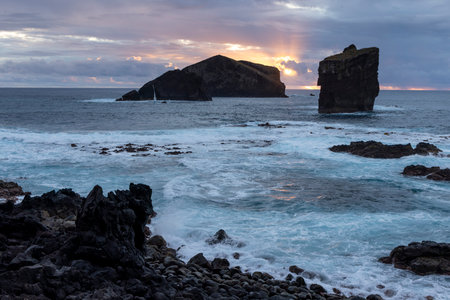 The ocean is calm and the sky is a beautiful orange color. The rocks on the shore are large and the water is blueの写真素材