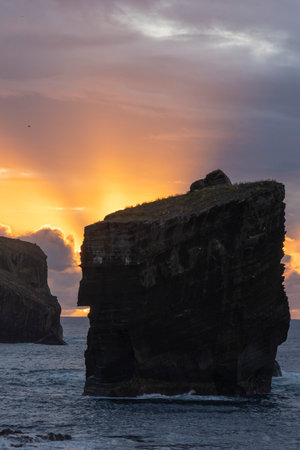 A large rock sits on the shore of a body of water, with the sun setting in the background. The scene is serene and peaceful, with the rock providing a sense of stability and strengthの写真素材