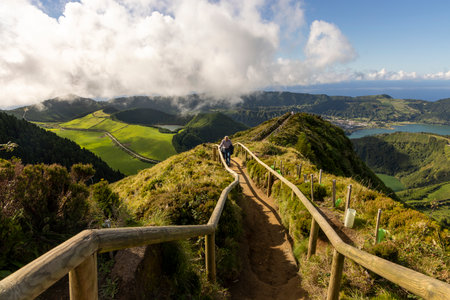 A man is walking on a path that leads to a mountain at Sete Cidades, Azores. The path is surrounded by trees and grass, and there is a view of the ocean in the distance. The sky is cloudyの写真素材