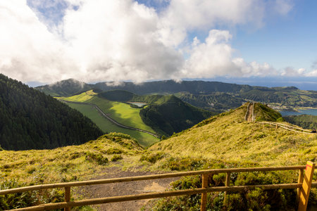 A beautiful view of a mountain range with a lush green field in the foreground at Sete Cidades, Azores. The sky is cloudy, but the sun is still shining through. The scene is peaceful and serene, with a sense of calmnessの写真素材