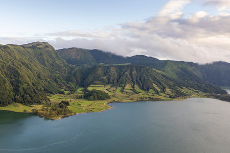 A beautiful mountain range with a lake in the foreground at Sete Cidades, Azores. The lake is surrounded by green hills and trees. The sky is clear and the sun is shiningの写真素材