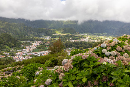 A mountain range with a town in the valley below. The town is surrounded by green hills and the mountains are covered in clouds at Furnas, Azoresの写真素材