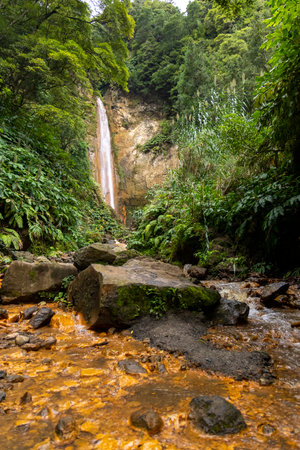 A waterfall is seen in the foreground of a lush green forest. The water is brown and murky, and the rocks surrounding the stream are large and jagged.の写真素材