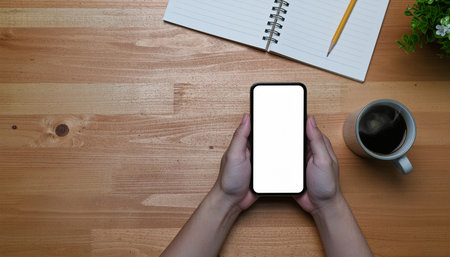 Mockup image of a woman's hands holding black mobile phone with blank white desktop screen on wooden tableの素材