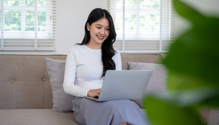 Happy asian woman using laptop on sofa in living room at homeの素材