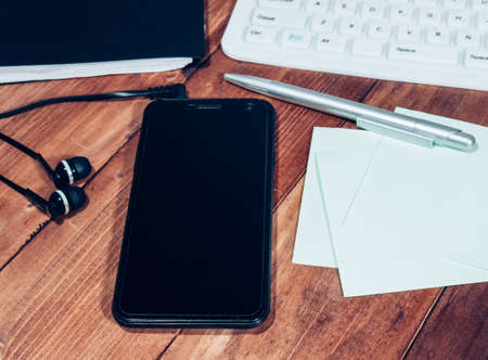 Workspace of a wooden table with a white keyboard, a black smartphone with headphones and a diaryの写真素材