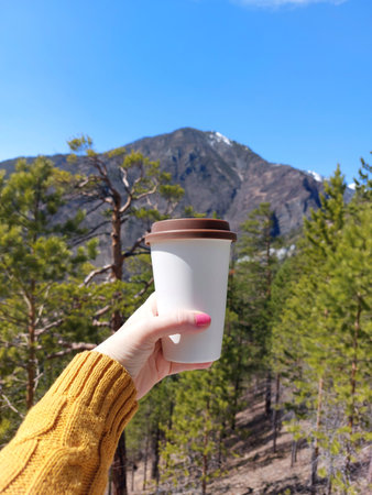 Female hand holding a paper cup of coffee on the background of the mountainsの写真素材