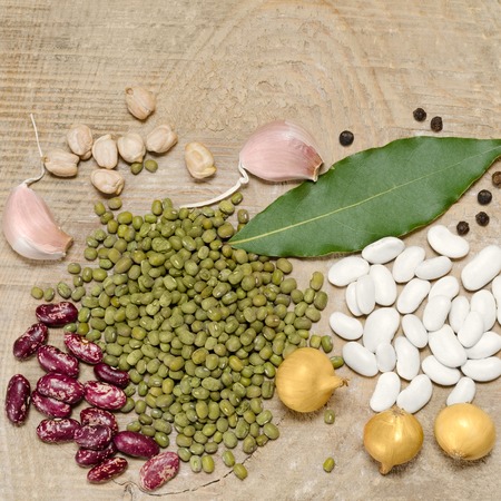 Different varieties of beans, onions, garlic and spices lie in heaps on the old wooden background.
