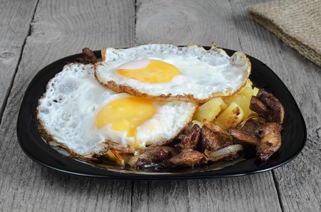 Fried eggs with potatoes and mushrooms on a black plate and a rough grey Boardの写真素材