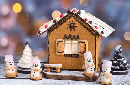 Christmas cake in the shape of the house, chocolate Christmas tree and sweet snowmen. On a wooden surface dusted with powdered sugar, colorful bokeh on the backgroundの写真素材