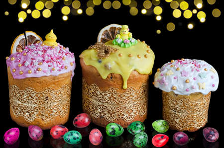 Decorated Easter cakes and colorful quail eggs on a black horizontal background with golden bokeh....