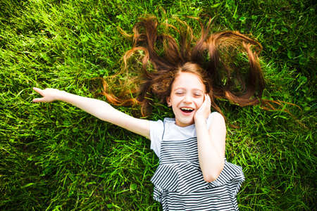 The girl lies on the grass laughing with her eyes closed and points with her finger to the side.