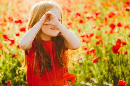 A small red-haired girl in a beautiful field looks at the camera through her fingers in the order. Simulating binoculars.の写真素材