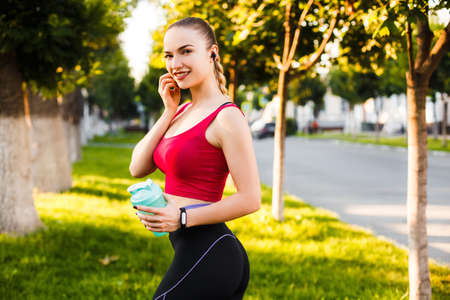 The girl listens to music in headphones and drinks protein from a shaker after a morning run in a summer city.の写真素材
