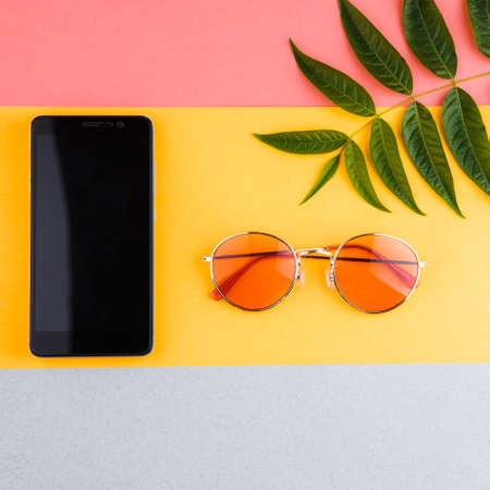 Composition of summer. A smartphone with sunglasses and a tropical plant on a colorful bright...
