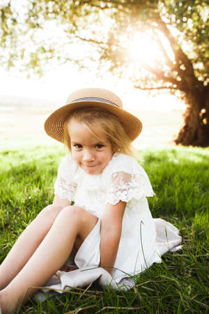 A cute baby girl in a straw hat and white dress is sitting on the grass.の写真素材
