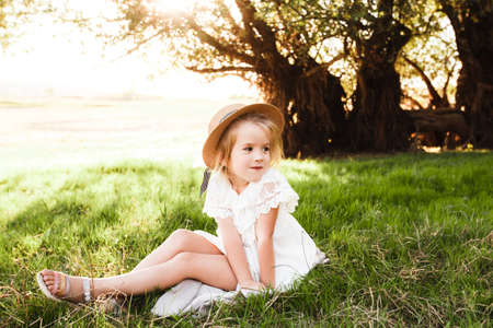 A cute baby girl in a straw hat and white dress is sitting on the grass.の写真素材