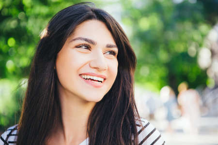 A brunette girl with a beautiful white smile against a green city background. Close-up. Space for text.の写真素材
