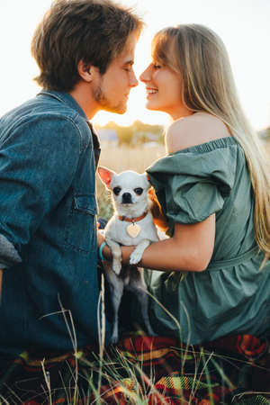 A young couple are sitting on a yellow autumn grass and hugging their white chihuahua breed dog.の写真素材