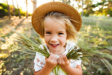 A cute baby girl in a straw hat and white dress is sitting on the grass.の写真素材