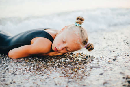 A teenage girl is sitting on the beach in a black swimsuit.