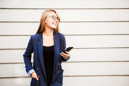 A blond girl in a blue jacket and glasses on the background of a light striped wall chatting in her smartphone.の写真素材