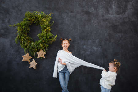 Funny children girls in warm sweaters against the background of a black wall with a Christmas wreath. Christmas concept, space for text.の写真素材