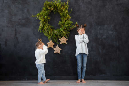 Funny children girls in warm sweaters against the background of a black wall with a Christmas wreath. Christmas concept, space for text.の写真素材
