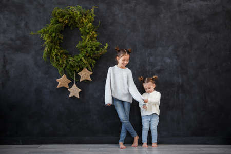 Funny children girls in warm sweaters against the background of a black wall with a Christmas wreath. Christmas concept, space for text.の写真素材
