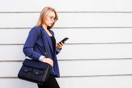 A blond girl in a blue jacket and glasses on the background of a light striped wall chatting in her smartphone.の写真素材