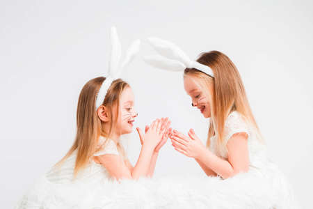 Little blonde twins in white dresses with rabbit ears. Studio photo on gray background. Kids...